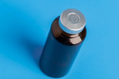 Kehribar rengi aşı enjeksiyon şişesinin seçici odak noktası olan bir makro çekim. Düz mavi arka plan üzerine kurulmuş..