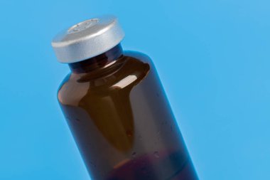 Kehribar rengi aşı enjeksiyon şişesinin seçici odak noktası olan bir makro çekim. Düz mavi arka plan üzerine kurulmuş..