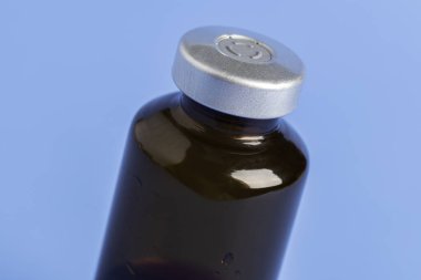 Kehribar rengi aşı enjeksiyon şişesinin seçici odak noktası olan bir makro çekim. Düz mavi arka plan üzerine kurulmuş..