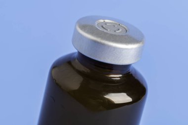 Kehribar rengi aşı enjeksiyon şişesinin seçici odak noktası olan bir makro çekim. Düz mavi arka plan üzerine kurulmuş..