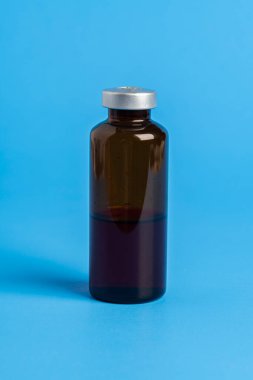 Kehribar rengi aşı enjeksiyon şişesinin seçici odak noktası olan bir makro çekim. Düz mavi arka plan üzerine kurulmuş..