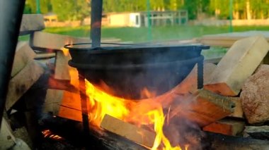 Kamp ateşi pişirme. Kürsüde büyük bir dökme demir kazan var. Ateşten gelen ateş kazanı ısıtır. Kırmızı alevler. Ateş yakmak için özel bir yer, şenlik ateşi. Kaya ve kiremit taşlarından yapılmış eskrim. - Rusya. İyi akşamlar. Yaz.