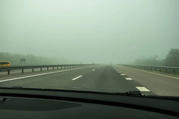 Siste asfalt yol. Arabanın ön camından yol manzarası var. Arabası olmayan yüksek hızlı bir pist. Yoğun sis görüş mesafesini kısıtlıyor. - Günaydın. Sis.