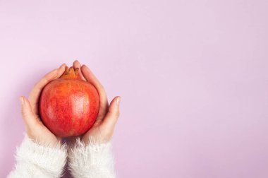 Beyaz pofuduk süveter giyen genç bir kızın elleri pembe arka plan fotokopi alanı üzerinde olgun bir nar tutuyor.