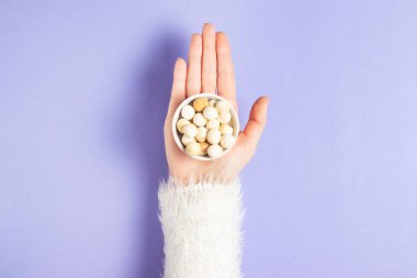 The palm of a young girl in a white fluffy sweater holds a plate with traditional dry kurut cheese on a purple background clipart