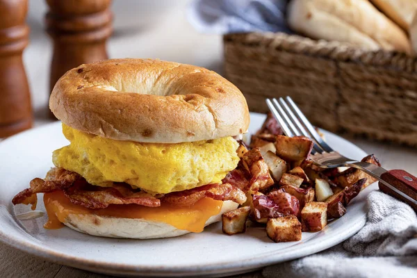 Breakfast sandwich with egg, bacon and cheese on a bagel with fried potatoes on a plate