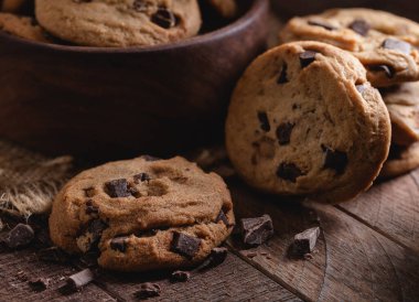 Çikolata parçalı kurabiyeler ve çikolata parçaları arka planda bir kase kurabiyeyle ahşap bir masada.