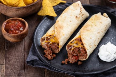 Biftekli burrito, kızarmış fasulye ve peynirli siyah tabakta salsa ve tortilla cipsi.
