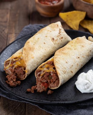 Biftekli burrito, kızarmış fasulye ve peynirli siyah tabakta salsa ve tortilla cipsi.