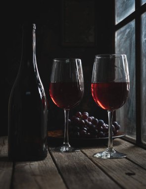 İki kadeh kırmızı şarap ve şişe, arka planda üzümler olan bir pencerenin yanında, ahşap bir masada.