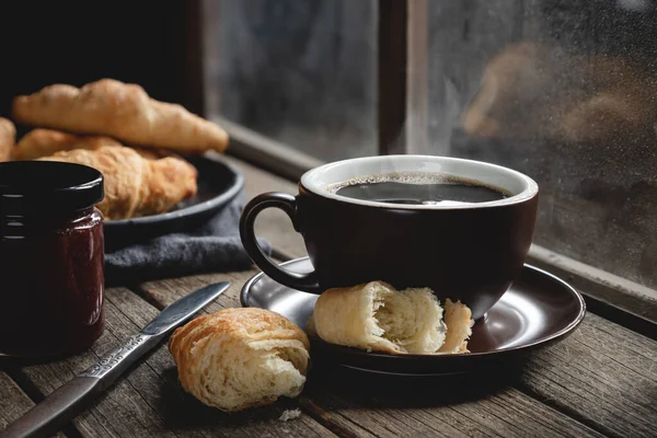 Hot cup of coffee with croissants on a rustic wooden table next to window