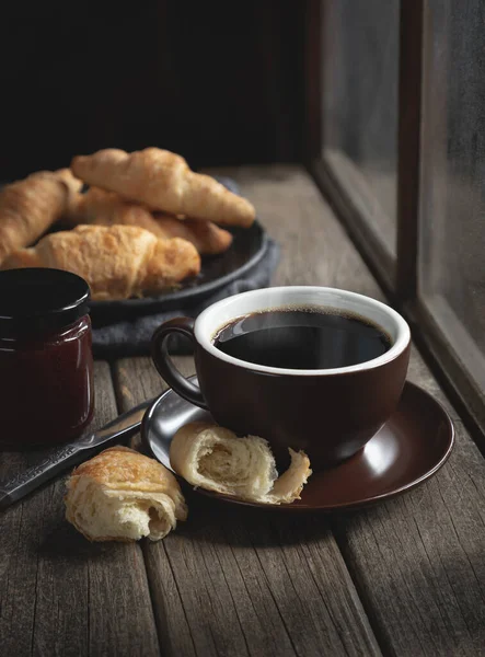 Hot cup of coffee with croissants on a rustic wooden table next to window
