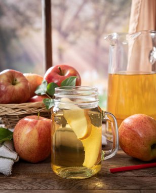 Bir bardak elma suyu ve elma, ahşap bir masanın üzerinde, arka plan manzaralı bir pencerenin yanında.