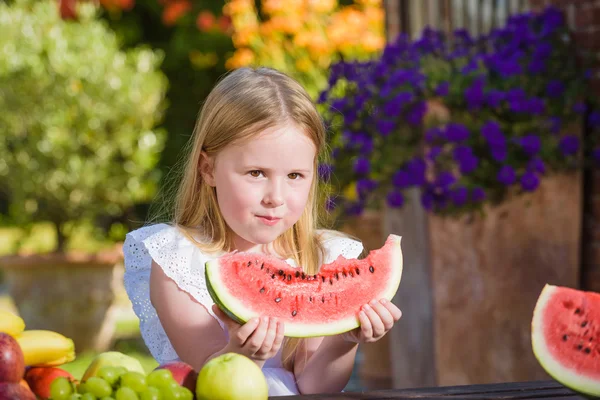Šťastná dívka s velkým červeným dílek melounu sedí na rustikální tabels v letní zahradě. — Stock fotografie