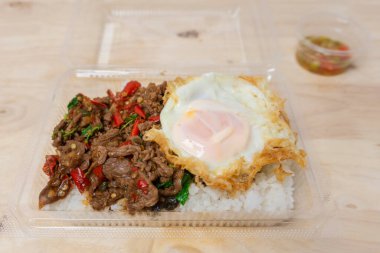 Üzerine kızarmış sığır eti ve ahşap masadaki plastik kutuda kutsal fesleğenle kaplanmış pirinç, popüler Tayland sokak yemekleri..