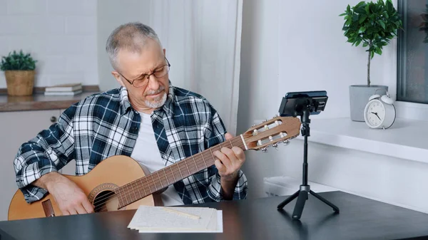 An elderly male blogger teaches guitar playing in an online lesson at home.