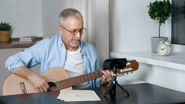 An elderly male blogger teaches guitar playing in an online lesson at home.