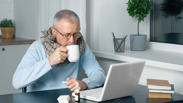 A sick elderly person sits at a laptop and suffers from the flu and a high fever.