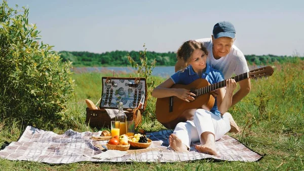 An elderly slender couple arranges a romantic picnic on the beach on a sunny summer day: a woman learns to play the guitar for her husband, and he teaches her to correctly take the chords on the