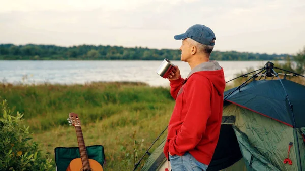 An elderly male tourist with an acoustic instrument enjoys nature and the sunset sunlight. People, lifestyle, the concept of rest in old age. An elderly slender man with a guitar near the river at