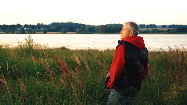 An elderly slender man enjoys nature and the evening landscape standing in the tall grass near the river at sunset. People, the concept of rest in old age.