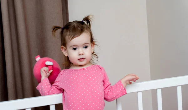 Симпатична дівчина стоїть на білому дитячому ліжечку і тримає рожеву іграшку з маленькими руками — стокове фото