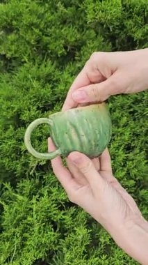 El yapımı Yeşil Seramik Kupa Lush Thuja 'ya karşı yavaşça el ele dönüyor. Yeşil seramik kupanın yakın çekim videosu her taraftan yavaşça dönüyor. Bardakta, organik dokusu ve parlak yeşil iç yapısı olan parlak benekli bir parlaklık var..