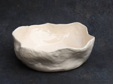 El yapımı bir seramik kasenin minimalist, düz olmayan kenarlı ve yumuşak desenli bir yüzeyi var. Fildişi parlaklık ışığı ince bir şekilde yansıtarak doğal kusurları ve heykel hacmini vurgular. Koyu desenli arka plan, yay