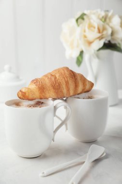 İki beyaz porselen kahve ve kruvasan. Arka planda bir buket narin krema çiçeği var. İki kişilik romantik bir sabah.