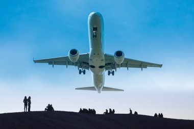 Mavi gökyüzüne karşı alçaktan uçan jet uçağı, yerde insan siluetleri.