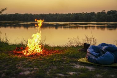 Bir uyku tulumu ateşin yanında yatıyor. Turistlerin ve balıkçıların nehir kıyısında uyuyacağı bir yer.