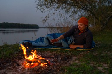 Uyku tulumunda bir turist nehir kıyısındaki bir yangında ısınıyor.