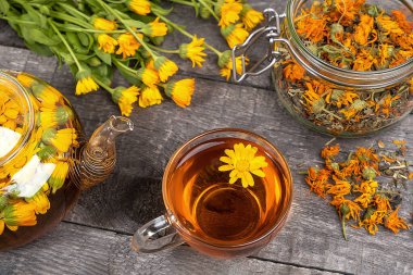 Bir fincan bitki çayı, şeffaf çaydanlık ve ahşap arka planda kadife çiçekleri. Calendula çayı sağlığınıza iyi gelir. Ön görünüm.