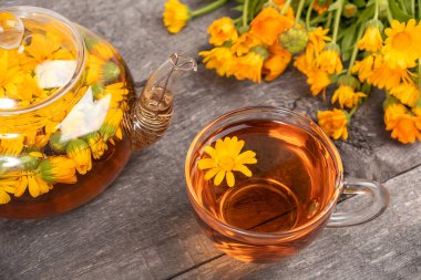 Bir fincan bitki çayı, şeffaf çaydanlık ve ahşap arka planda kadife çiçekleri. Calendula Çayı Sağlığınıza Yararlıdır.