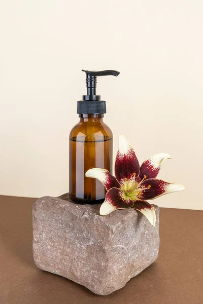 Brown glass bottle with pump of cosmetic products and lily flower on stone, beige and brown background. Natural Organic Spa Beauty Cosmetic concept Mockup.