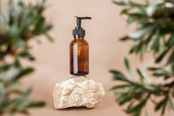 Brown glass bottle with pump of cosmetic products on stone framed by green leaves of branches, beige background. Natural Organic Spa Cosmetic Beauty Concept. Front view Mock up.
