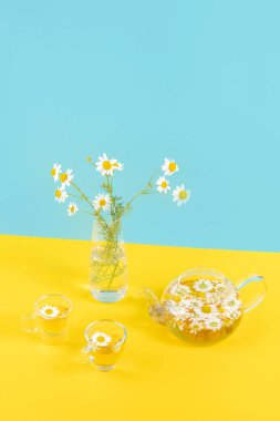 İki fincan papatya çayı şeffaf çaydanlık ve vazo mavi sarı arka planda papatya benzeri çiçekler. Papatya çayı sağlığınıza iyi gelir.
