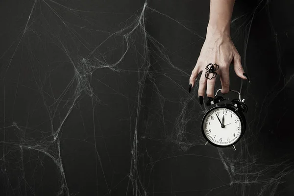 Hand mit langen schwarzen Nägeln und Spinnenring hält einen Wecker auf schwarzem Hintergrund mit Spinnweben. Glückliches Halloween-Urlaubskonzept — Stockfoto