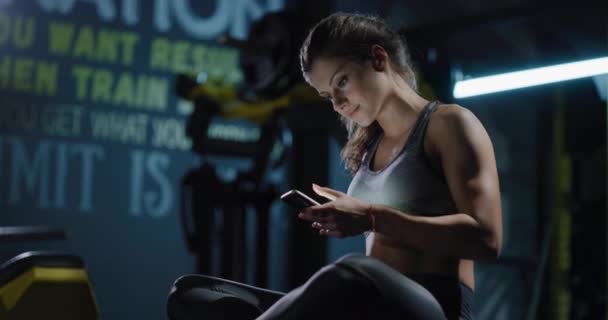 Молодая женщина в спортивной одежде держа смартфон смс в помещении фитнес-тренировки отдыха фитнес-технологии Концепция 4k — стоковое видео