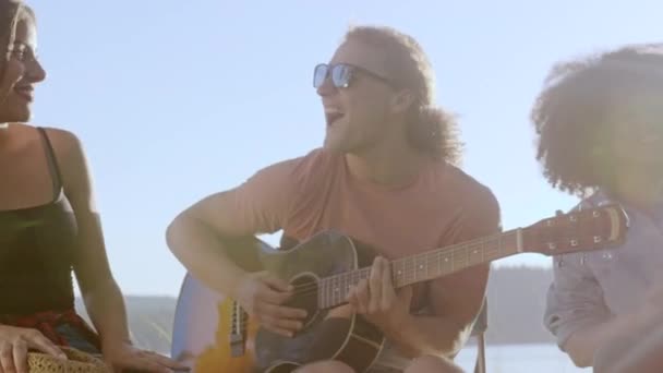 Grupo de amigos felizes tocar guitarra e sentar ao redor floresta acampamento fogo bater bebida rir e brincar música e felicidade na felicidade selvagem na natureza conceito câmera lenta tiro no épico vermelho w 8k. — Vídeo de Stock