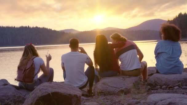 湖の海岸に座っている若い友人の魅力的な多人種グループ美しい夕日を見て社交親しみと観光旅行パーティーのコンセプトスローモーションショットレッドエピックW 8k — ストック動画