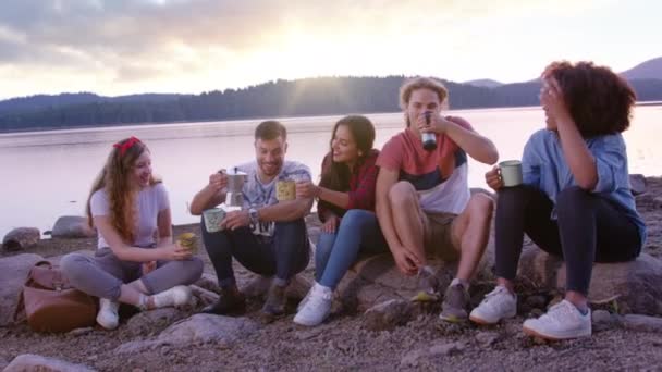 Różnorodna grupa atrakcyjnych młodych ludzi siedzieć i śmiać się na brzegu jeziora piękny wschód słońca rano pić kawę wakacje w koncepcji przyrody slow motion shot na czerwonym epic w 8k. — Wideo stockowe