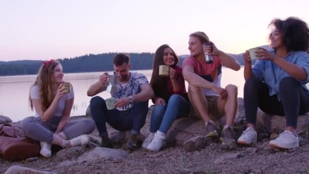 Olika grupp av unga män och kvinnor omfamna och skratta när du sitter vid sjön Shore tidig morgon dricka kaffe vänskap och lycka i den vilda picknick part i naturen koncept Slow Motion — Stockvideo