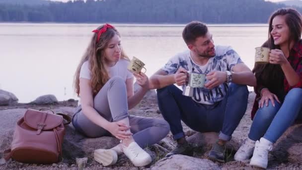 Feliz joven grupo de amigos abrazo y sentado en la orilla del lago compartir café naturaleza exploración turismo ocio concepto cámara lenta disparo en rojo épica w 8k — Vídeo de stock