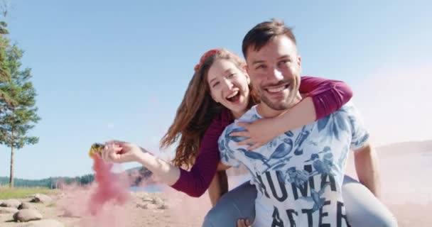 Szczęśliwy para mężczyzna nosić kobieta na jego plecach na plaży jezioro przechodzą przez chmury dym słoneczny dzień lato romantyczne wakacje młody miłość koncepcja slow motion strzał na czerwony epic w 8k. — Wideo stockowe