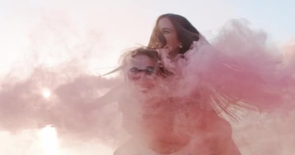 Atraente jovem feliz casal homem e mulher girando e rindo no piggyback no lago Shore passando através de nuvens de fumaça romântica fuga casamento e amor conceito lento movimento tiro no vermelho épico w — Vídeo de Stock