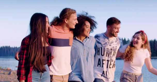 Grupo diverso de homens e mulheres jovens abraçam e riem enquanto caminham na praia do lago saltam férias na natureza conceito de festa de viagem câmera lenta tiro no épico vermelho w 8k. — Vídeo de Stock