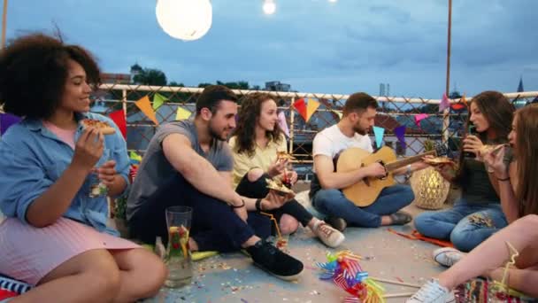 Gruppo di Diversi Teen Friends On Rooftop Sitting Down and Moving In Rhythm To Man Playing Guitar Happy Drinking And Eating Pizza Outdoor Festive Music Pizza Party Young Carefree Friendship Concept At — Video Stock