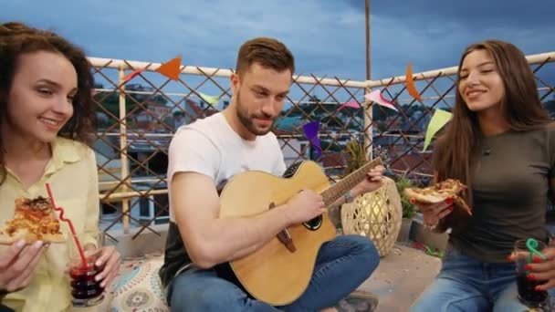 Zróżnicowane młodych ludzi Party na dachu siedzi i porusza się w rytm do człowieka gra na gitarze picie koktajli i jedzenia pizzy na świeżym powietrzu Uroczysta muzyka Pizza Party Beztroska przyjaźń Wesoła muzyka — Wideo stockowe