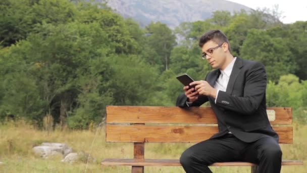 Jovem empresário feliz usando tablet sentado banco do parque — Vídeo de Stock